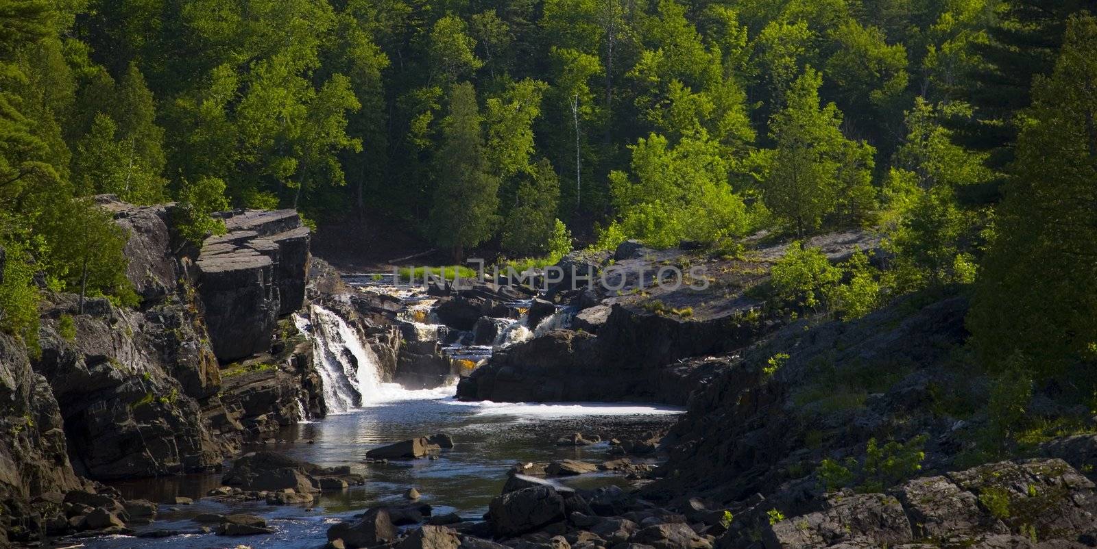 Water steps and falls down stone from a green forest into a blue river in Northern Minnesota.