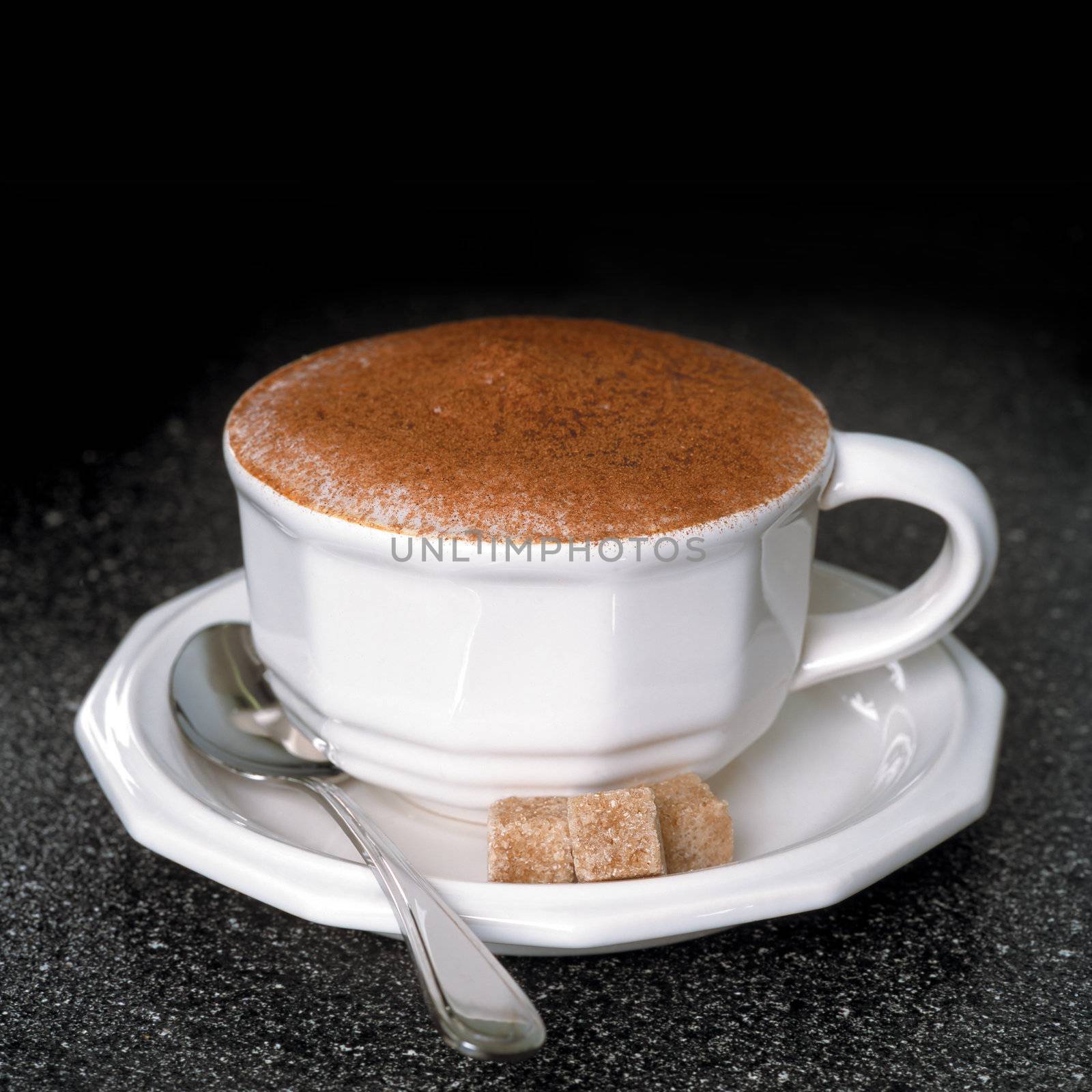 Hot cappuccino with spoon and sugar cubes on a dark wood table.

