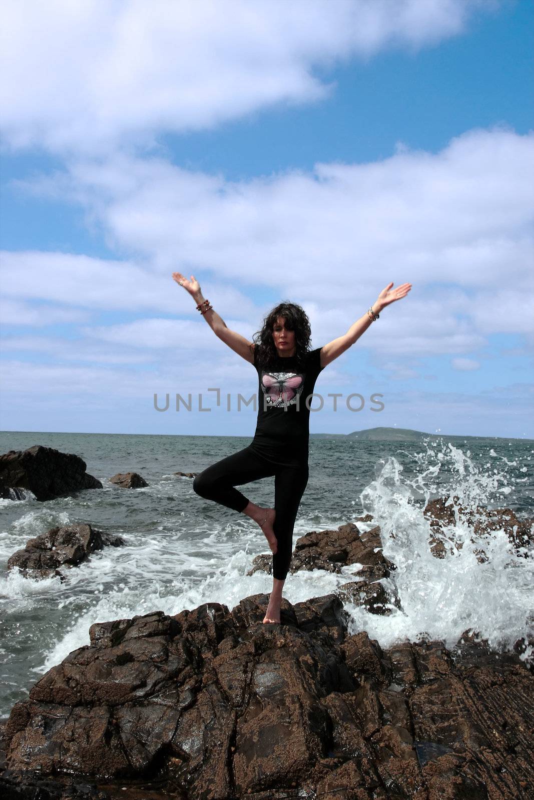 a beautiful woman doing yoga on the edge of the rocks with waves splashing to show a healthy way to live a happy and relaxed lifestyle in a world full of stress