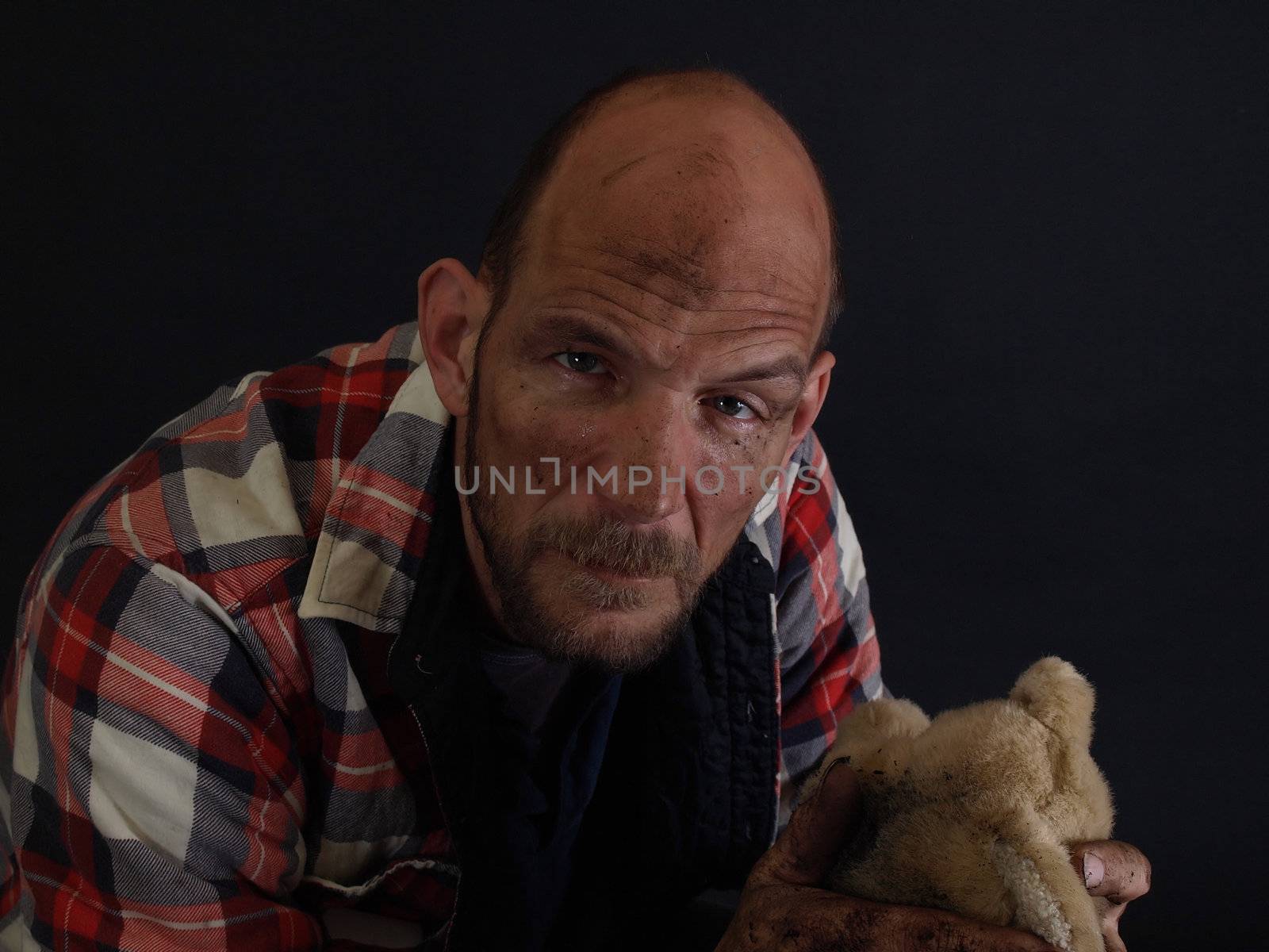 A dirt covered man holds an orphaned dirty stuffed teddy bear in one hand, a look of sadness on his face. Over a black background.