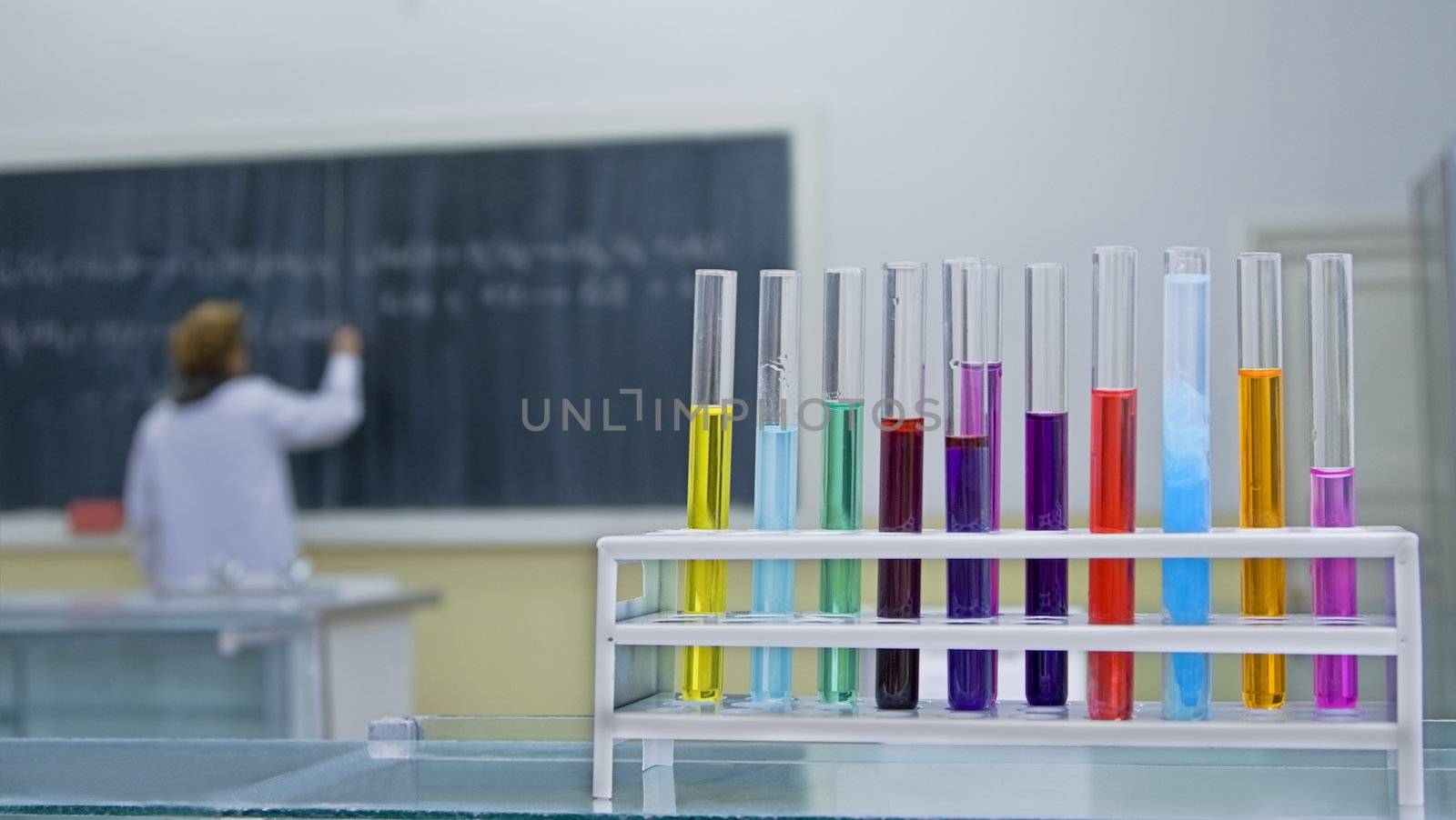 Image in a school chemiostry lab.Selective focus on the tubes in foreground.In the background there is a student writing someting on the blackboard.