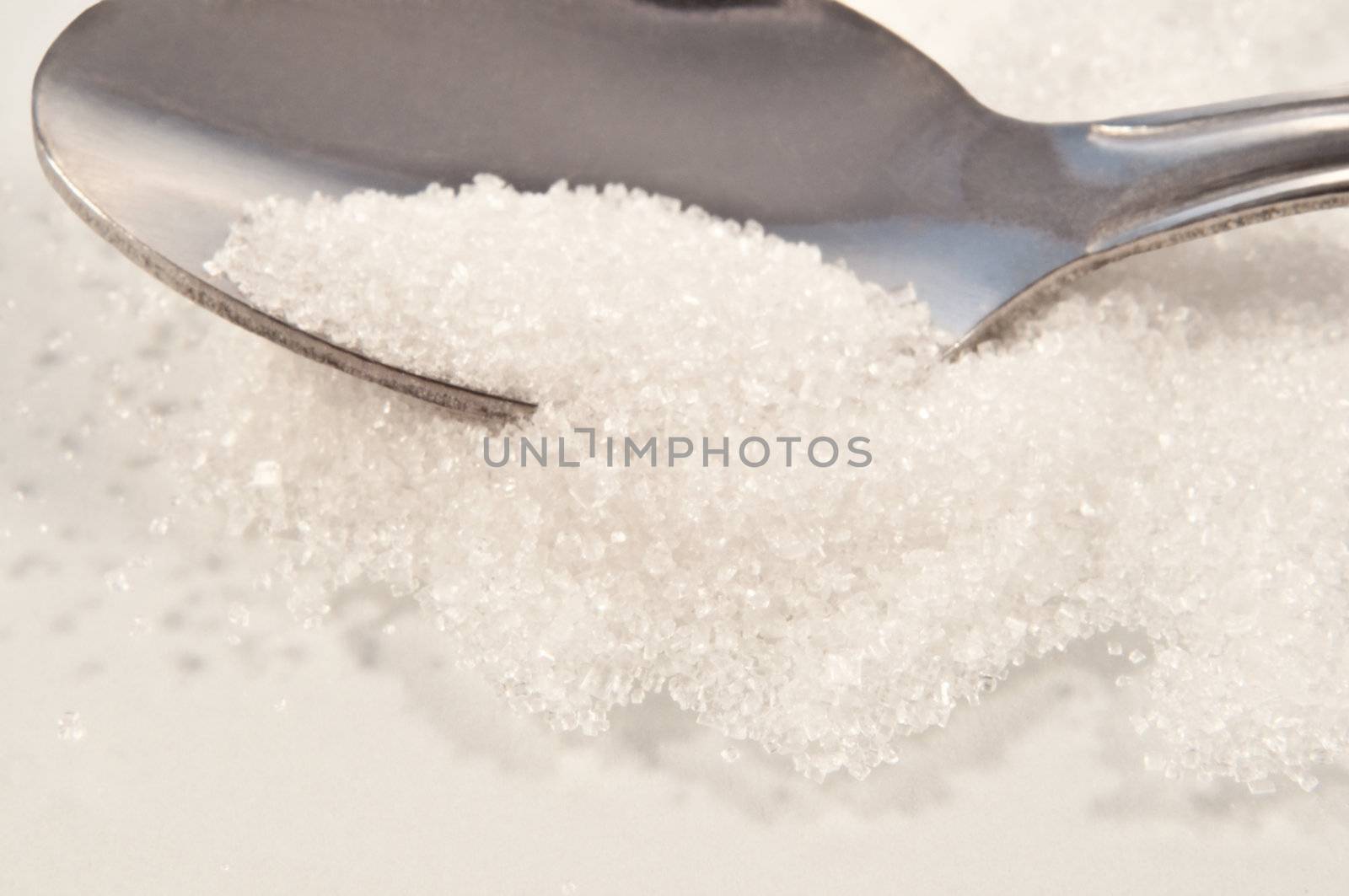 Close and low level capturing white granulated sugar spilling from a silver spoon onto a white reflective surface.