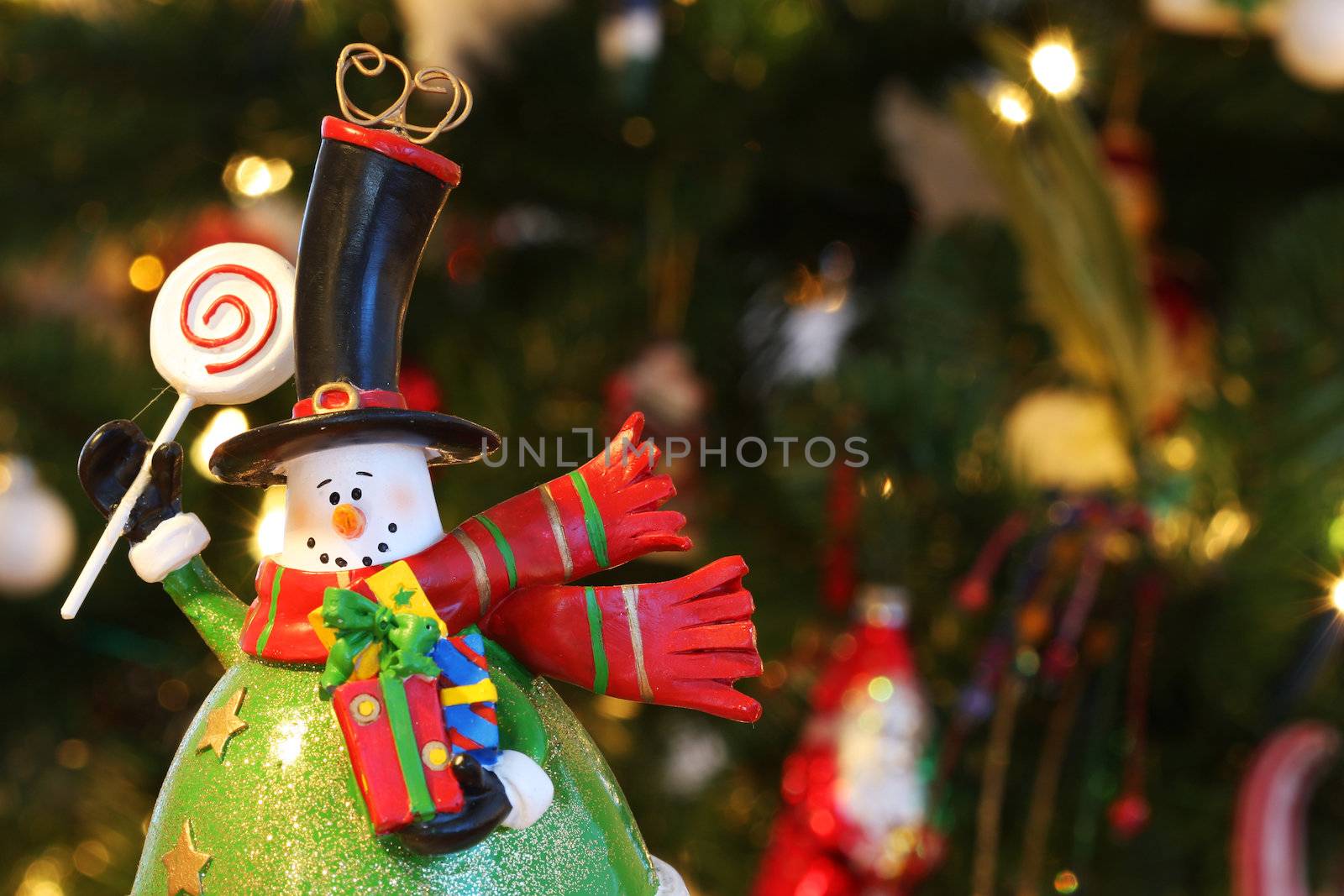 A christmas decoration in the form of a snowman, waving and holding christmas presents, decorated christmastree in background