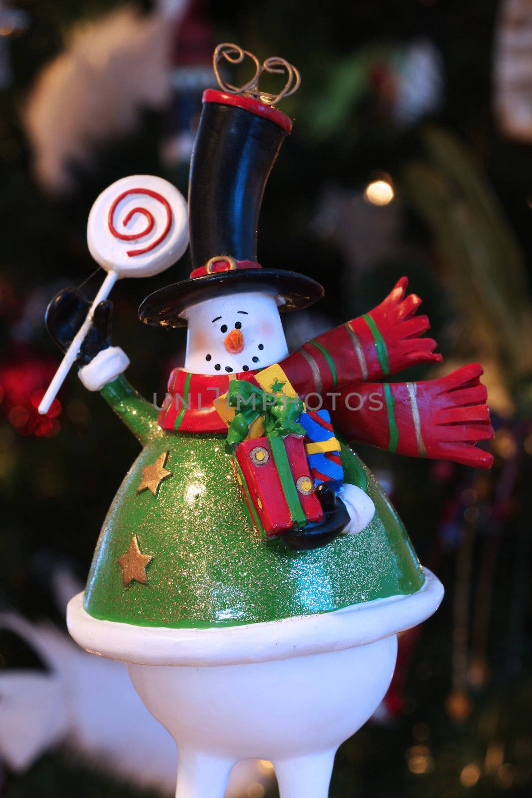 A decorative christmas snowman, holding gifts and waving, very blurry christmas tree in background