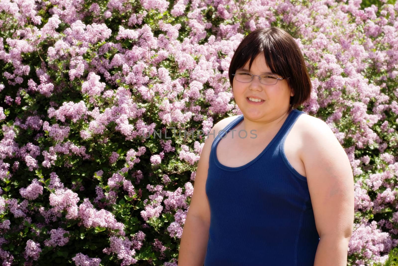 A young girl wearing a tank top standing in front of a blooming lilac bush