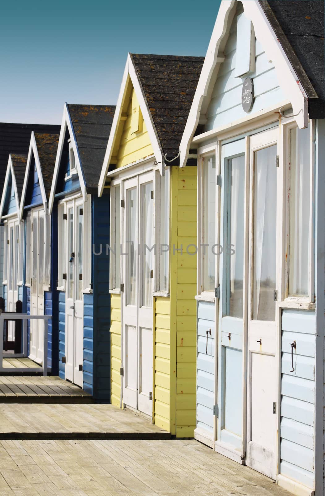 Front view of a row of wooden beach huts with wooden terraces located in Christchurch Dorset Hampshire UK. Huts painted in pale blue and yellow colours.