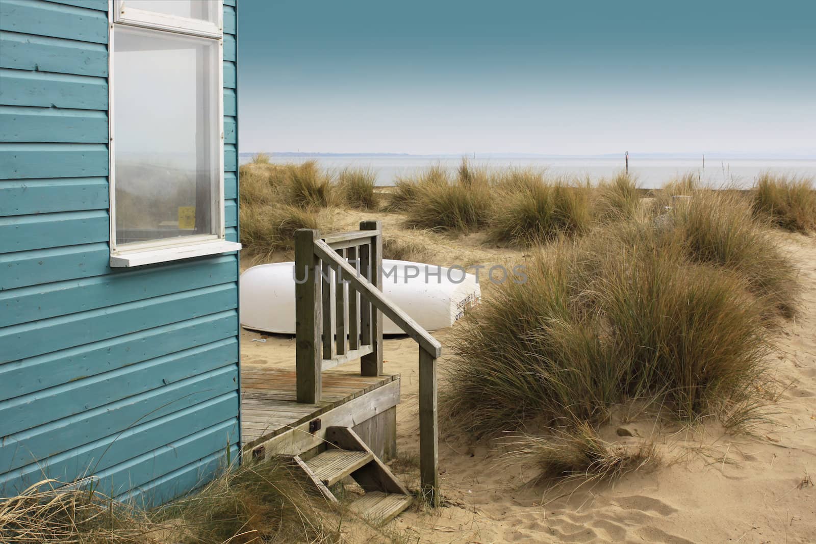 A view of a blue painted wooden beach hut with wooden terrace, looking towards the beach. An upturned white boat to front of beach hut rests amongst sand dunes. Set on a landscape format.