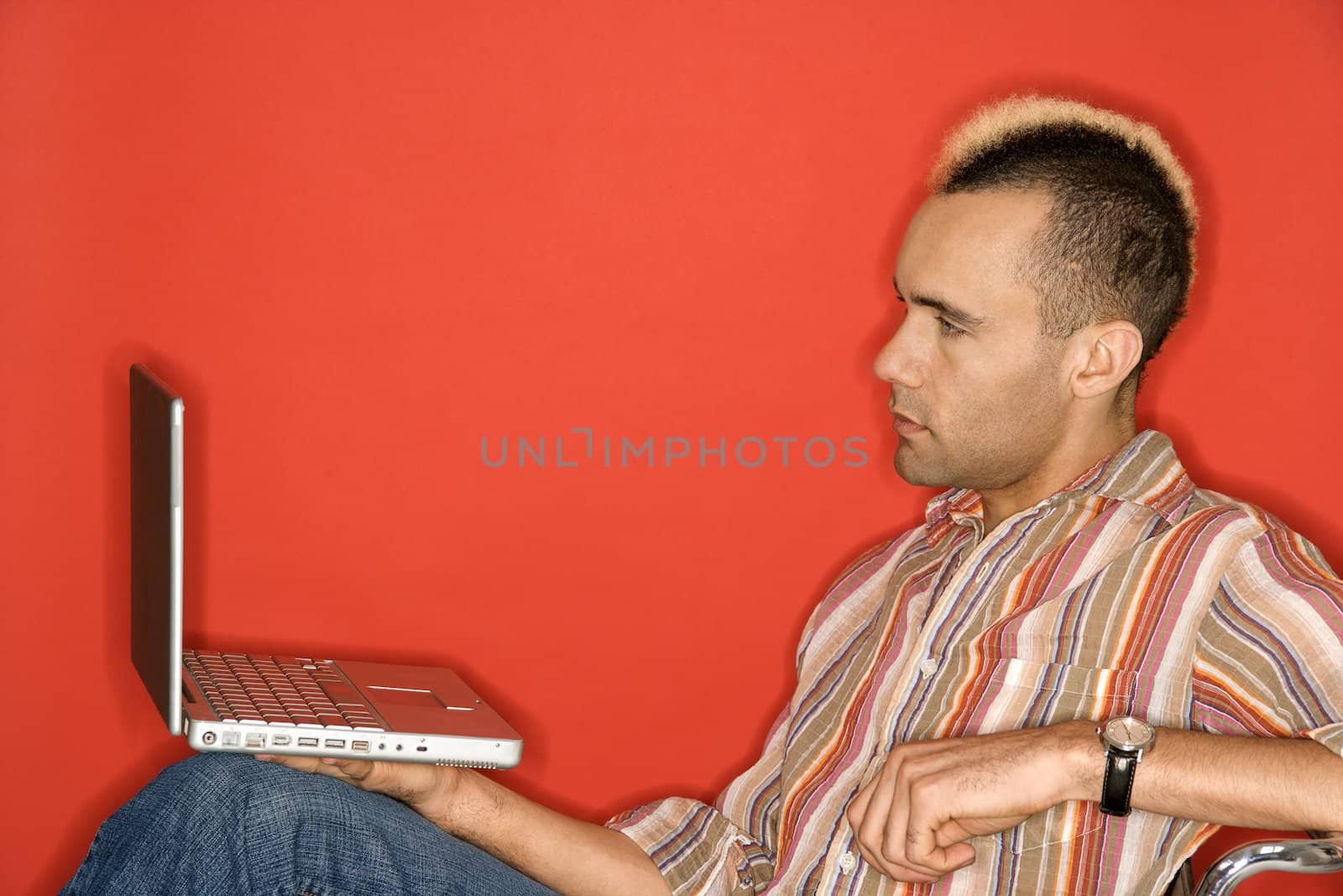 Caucasian man with mohawk holding laptop against red background.