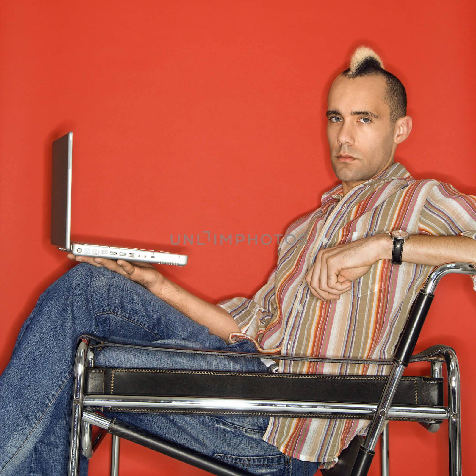 Caucasian man with mohawk holding laptop against red background.