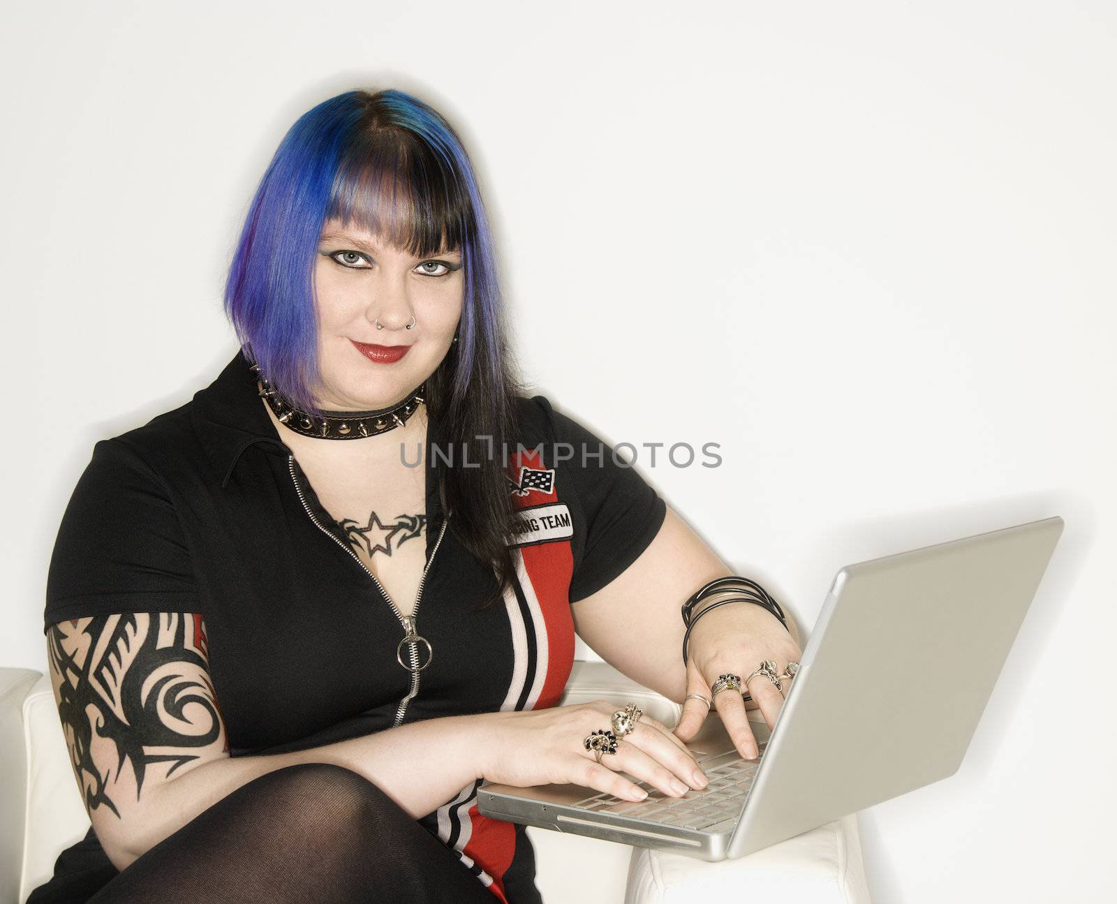 Portrait of Caucasian woman with blue hair, tattoo, and spike collar typing on laptop against white background.
