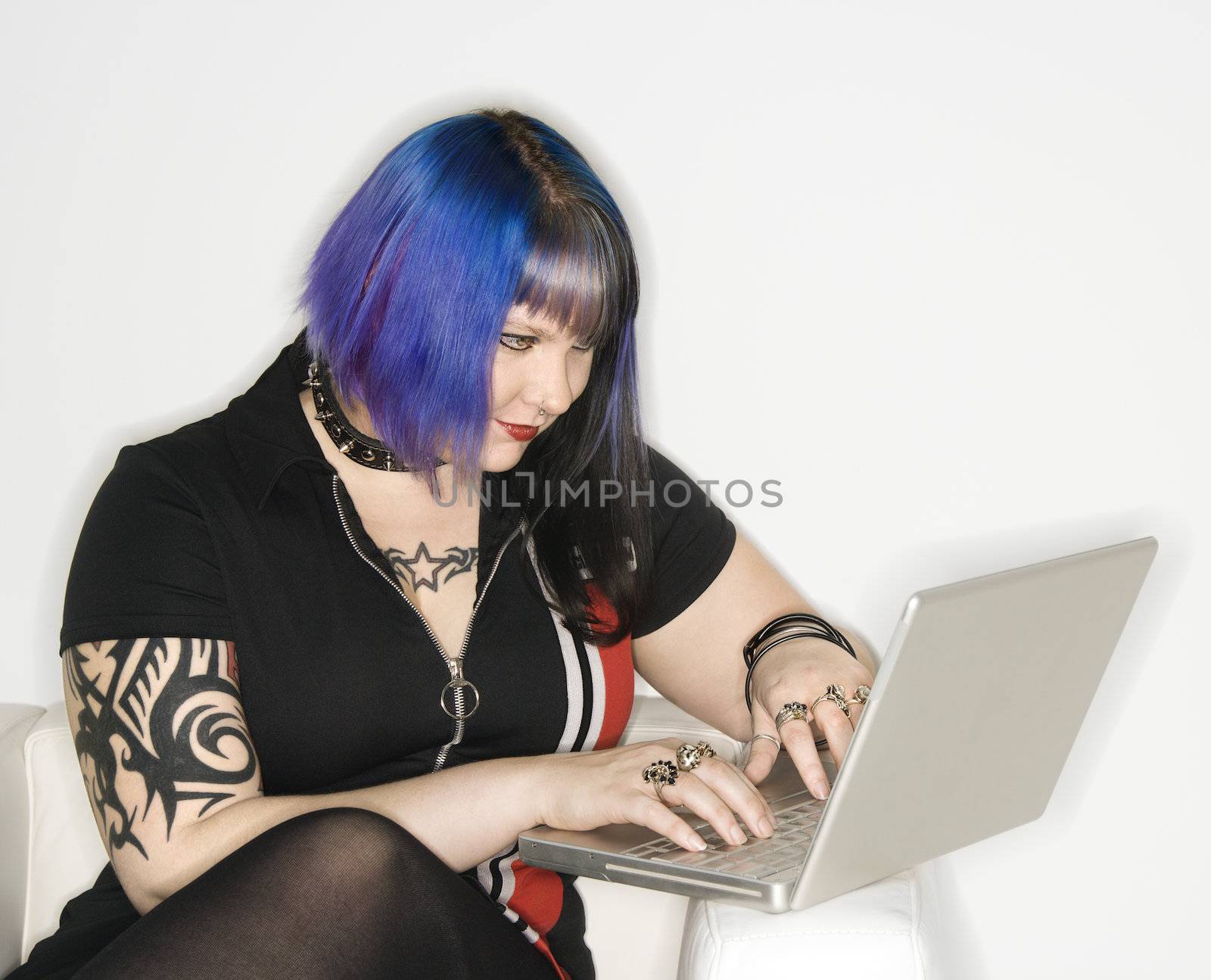 Portrait of Caucasian woman with blue hair, tattoo, and spike collar typing on laptop against white background.