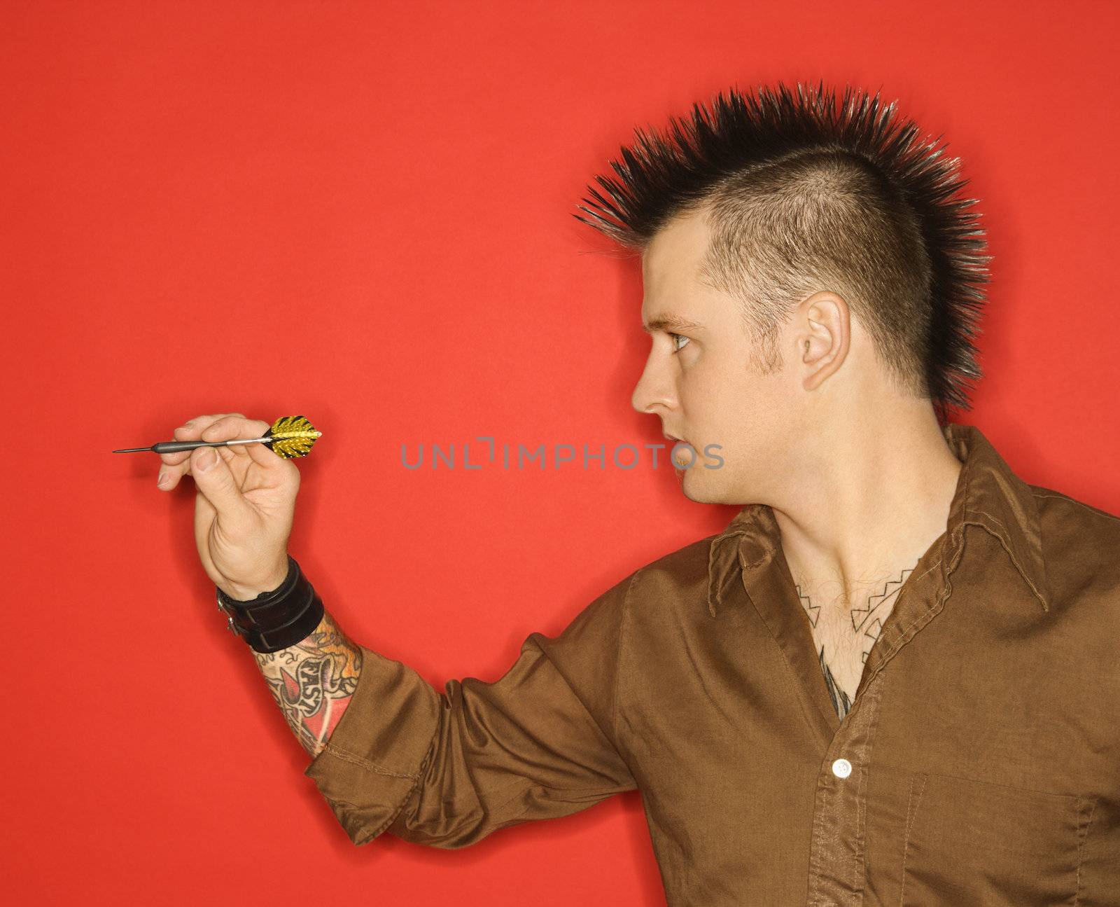 Side view of Caucasian man with mohawk holding dart against red background.