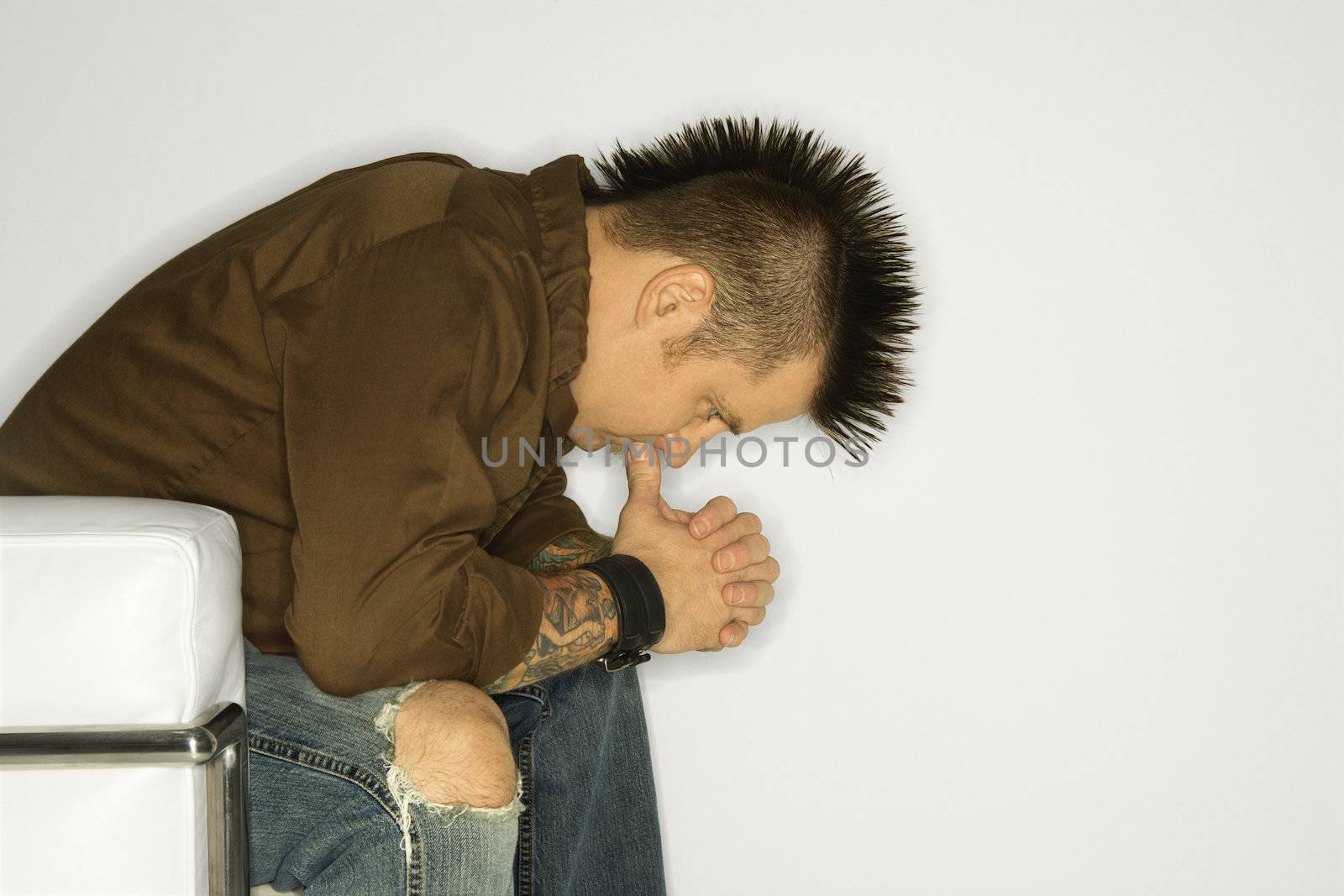 Side view of Caucasian man with mohawk sitting with hands to face against white background.