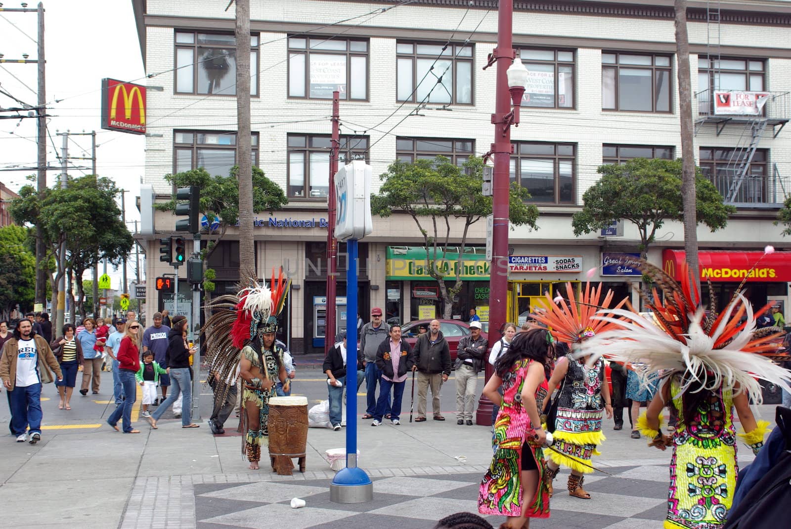 An Aztec street performers in full ceremonial dress plays a drum for women dancers on a street corner in San Francisco California.