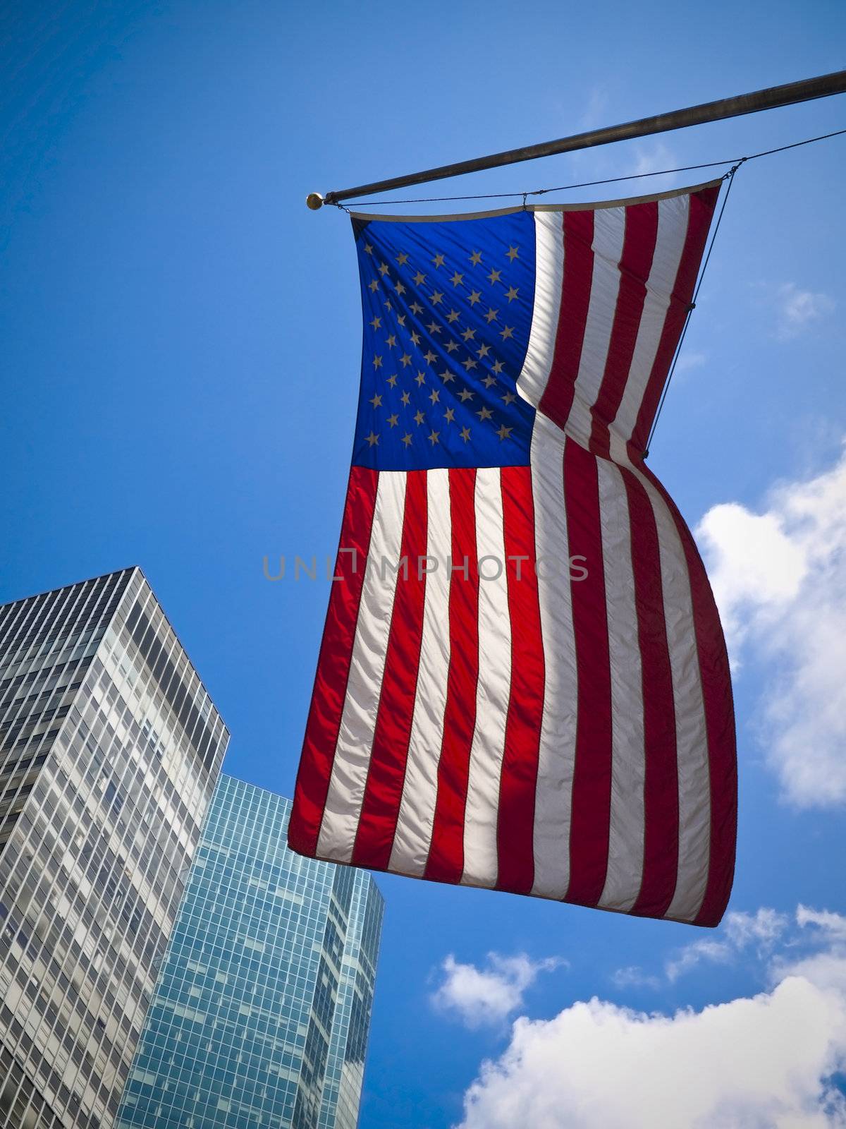 An American flag waving against two skyscrapers and a blue sky.