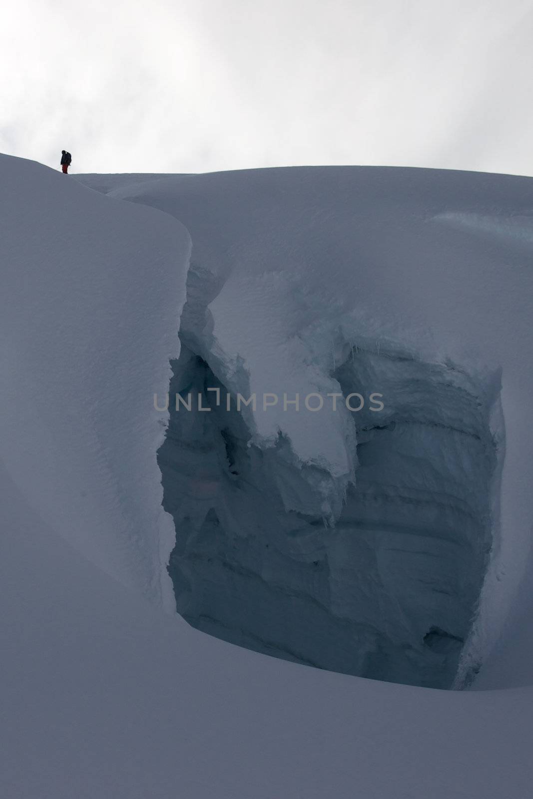 Side view of crevasse with tiny figure of a climber crossing it via a snow bridge