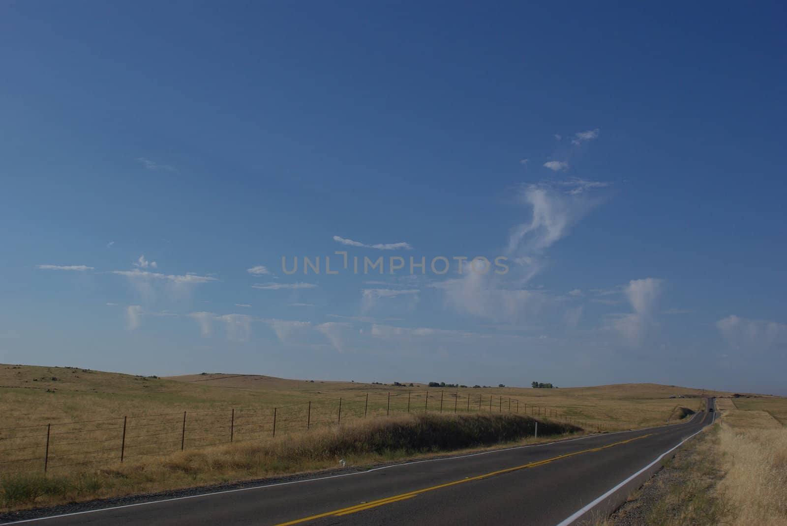 A 2 lane road cuts across prairie hills covered with dry grass with unusuale cloud formations in the background.