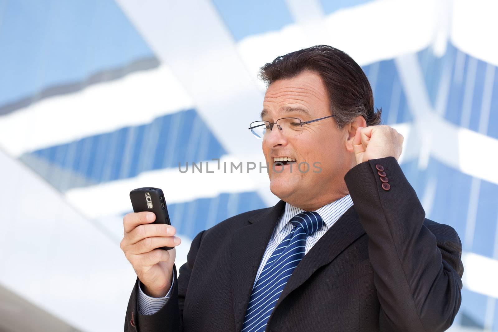 Excited Businessman Looking at Cell Phone Clinches His Fist in Joy Outside of Corporate Building.