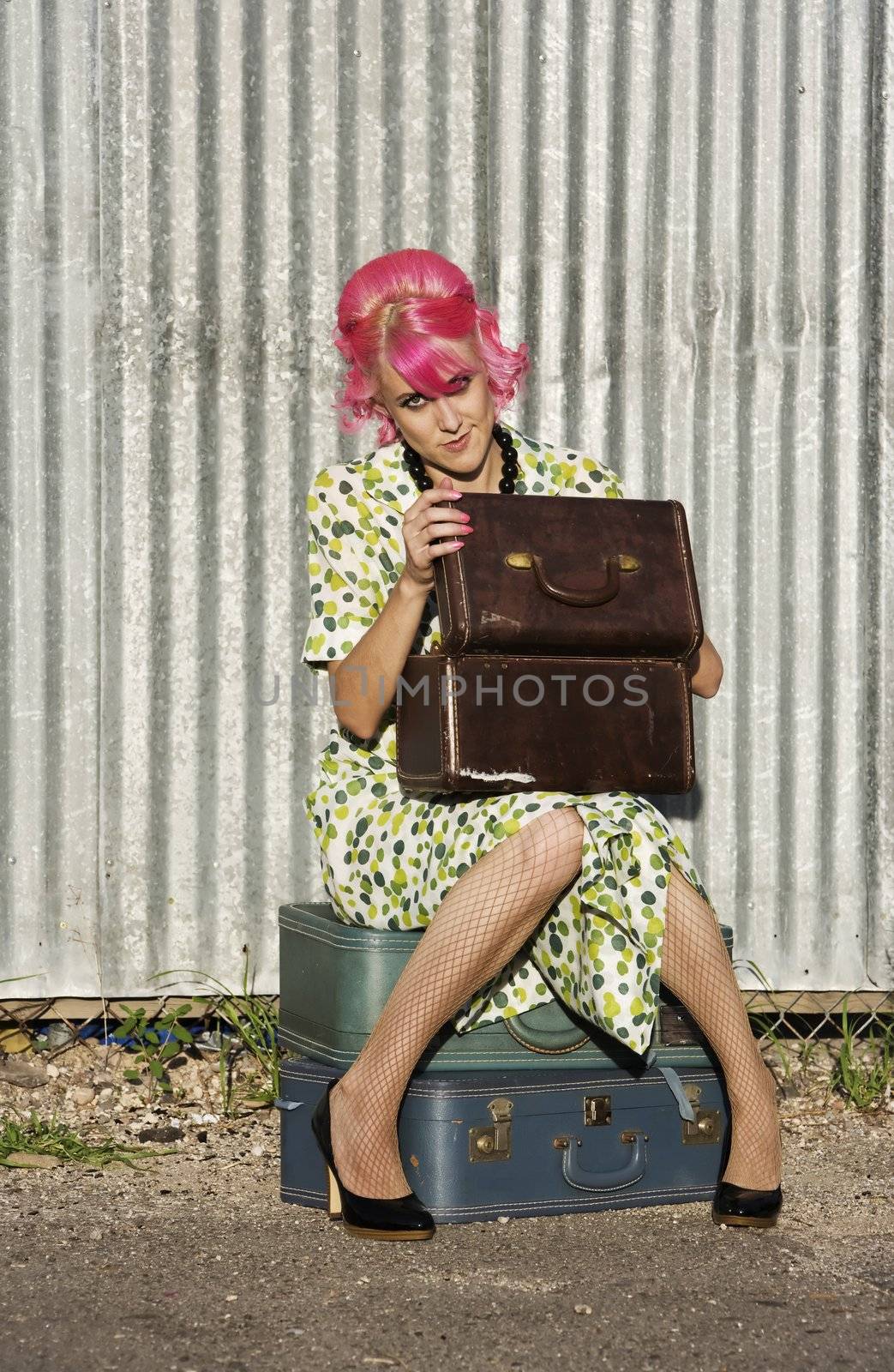 Woman with pink hair wearing polka dot dress in alley with suitcases