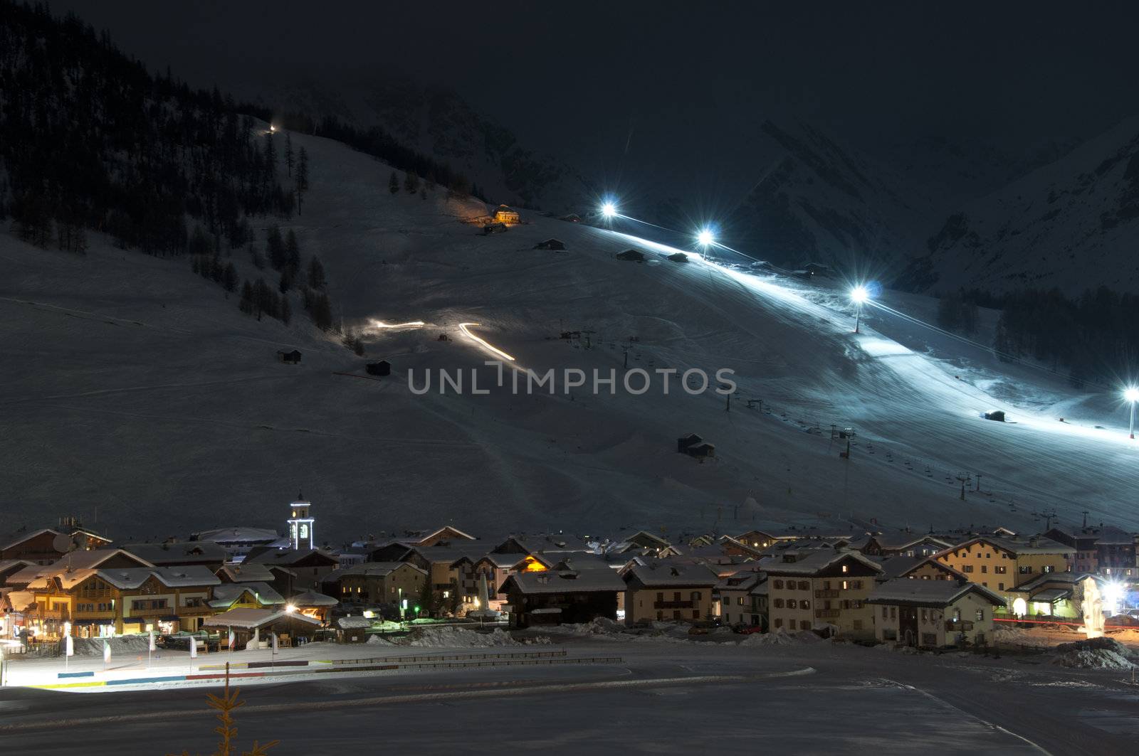 Ski village at night with slope lights, cross-country ski run, buildings, ice monument - shot in Livigno, Italian Alps