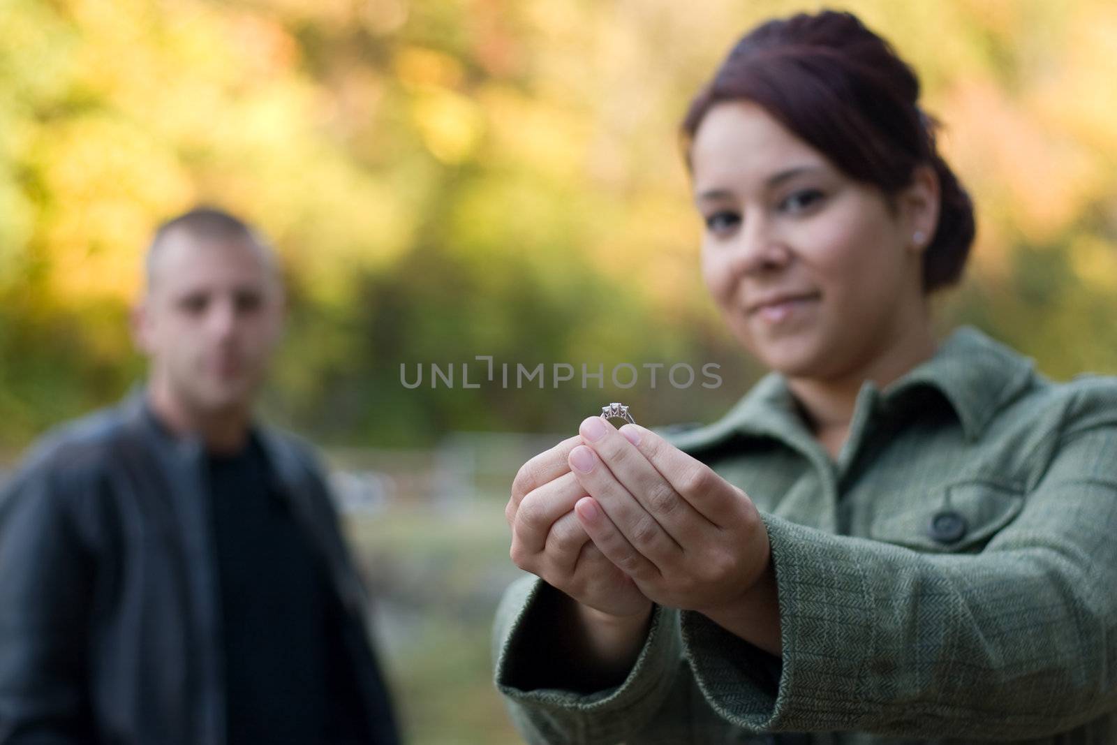 A young woman holding a diamond engagement ring with her fiance in the background.  Shallow depth of field with focus on the ring.