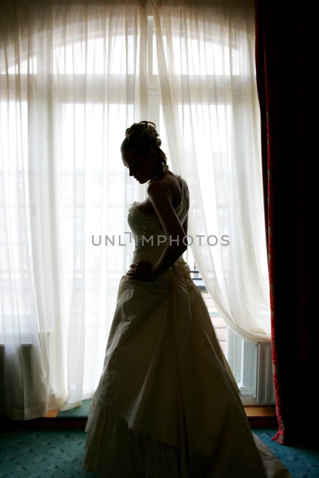 Silhouette of a beautiful bride in a traditional white wedding dress, stood by window.