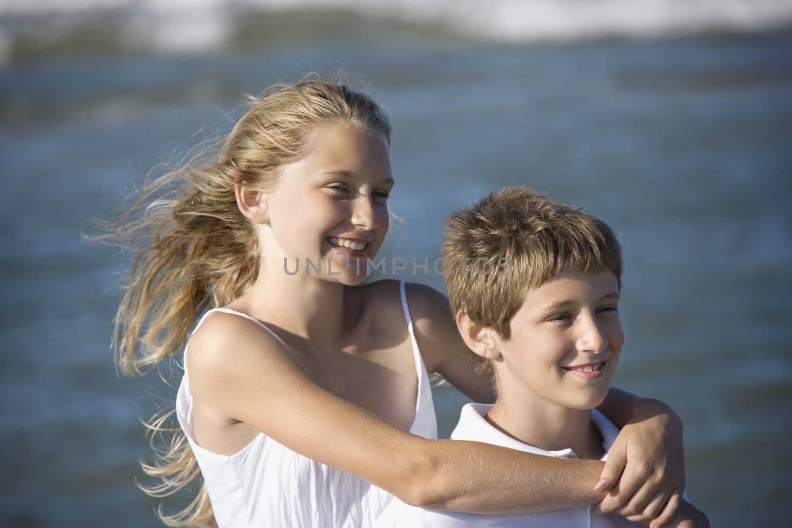 Caucasian pre-teen girl with arms around pre-teen boy on beach.