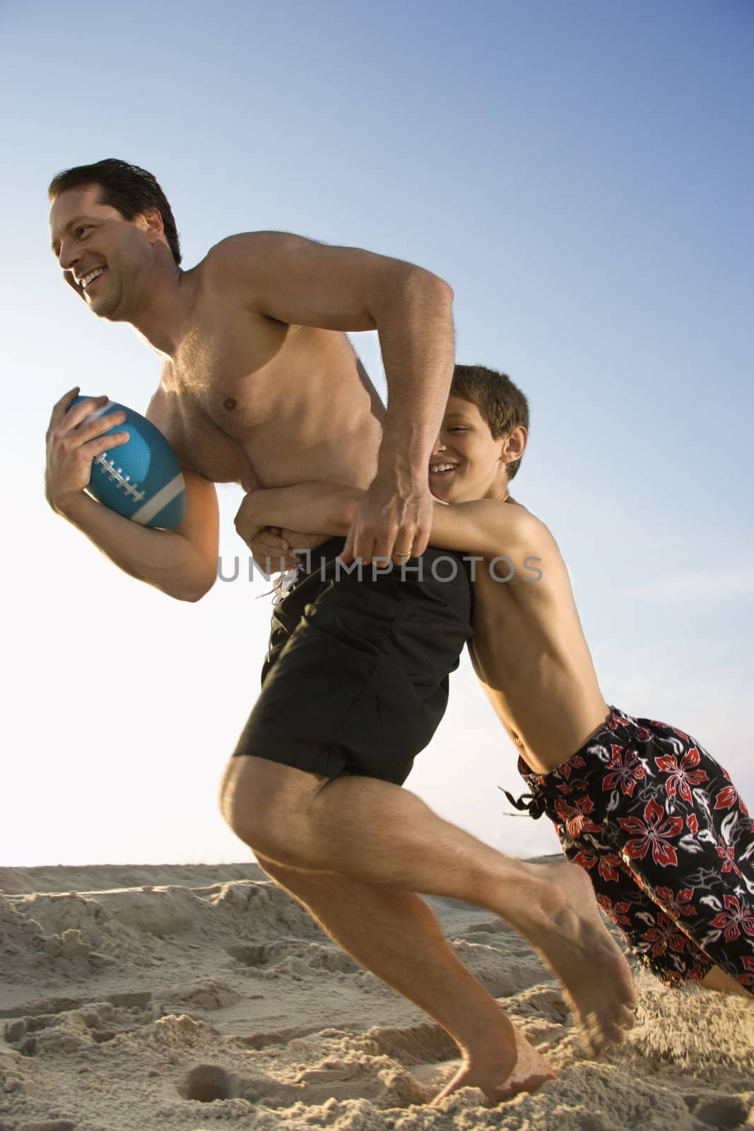 Caucasian mid-adult man running with football being tackled by pre-teen boy.