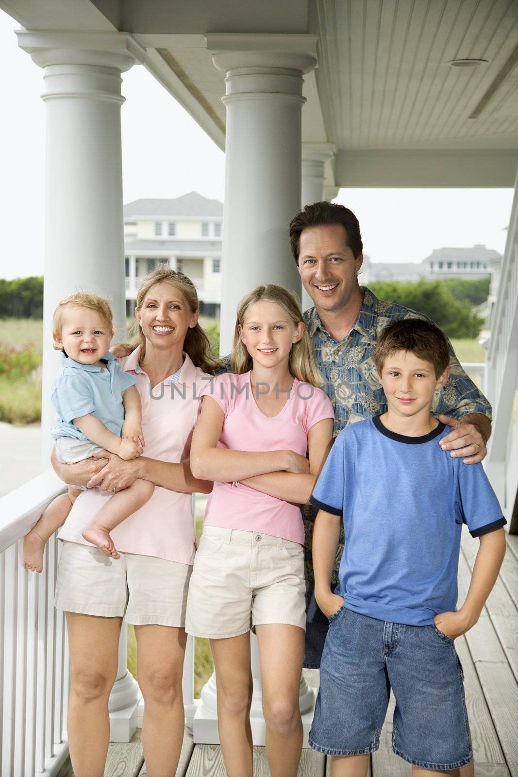 Family portrait of Caucasian mid-adult man and woman with pre-teen girl and boy and male toddler, standing on porch.