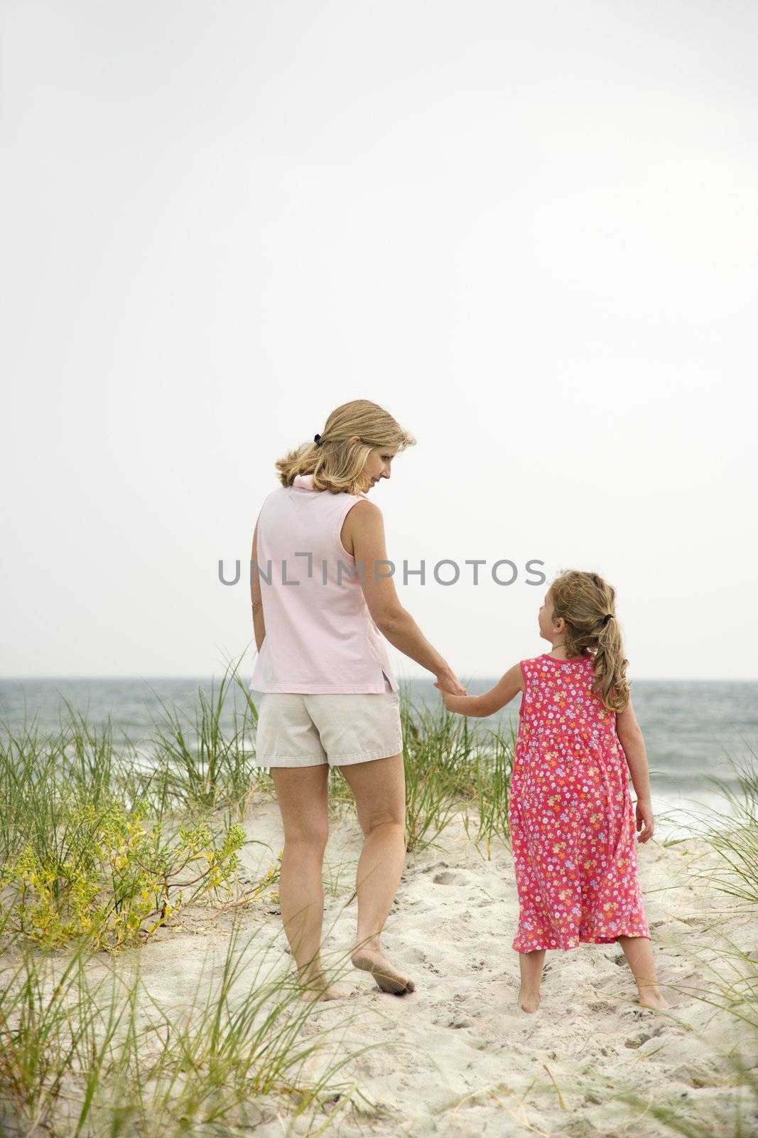 Caucasian mid-adult woman walking and holding hands with Caucasian female child on beach.