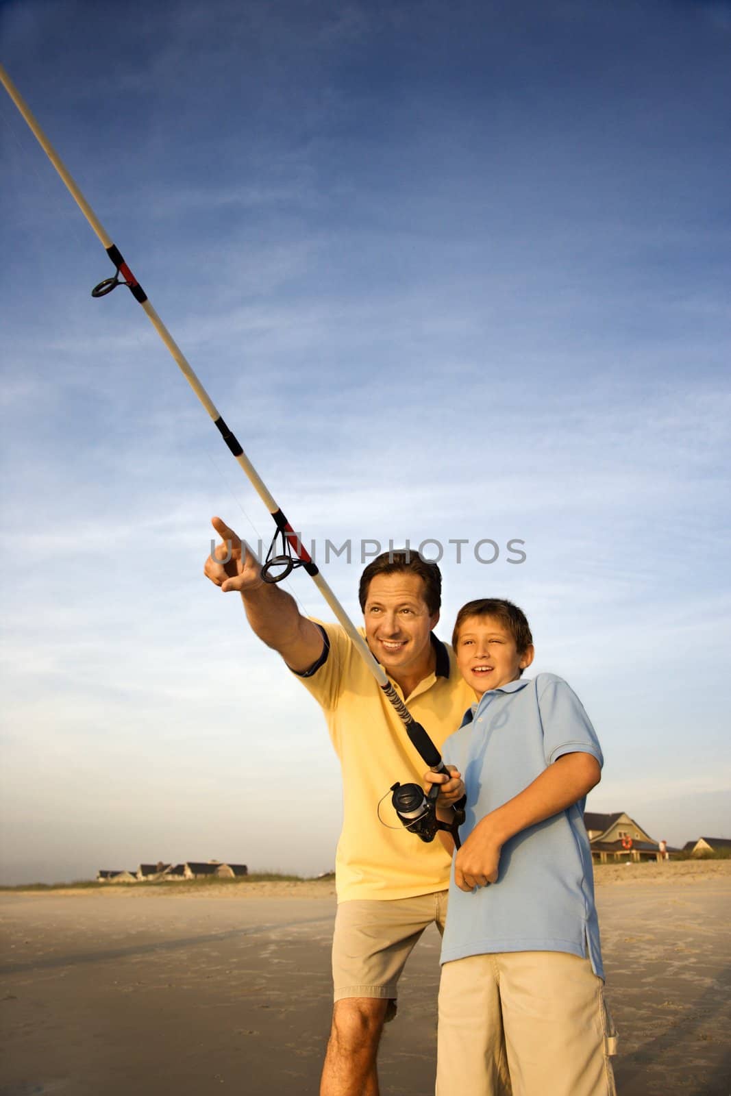 Caucasian mid-adult man shore fishing on beach with pre-teen boy and pointing.