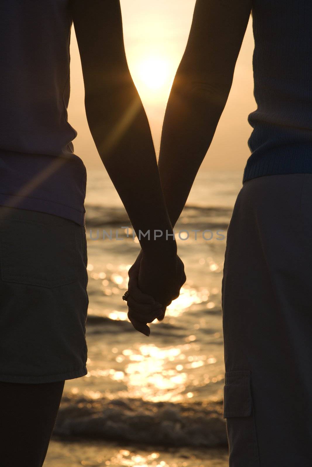 Caucasian mid-adult mother and teenage daughter standing on beach at sunset holding hands.