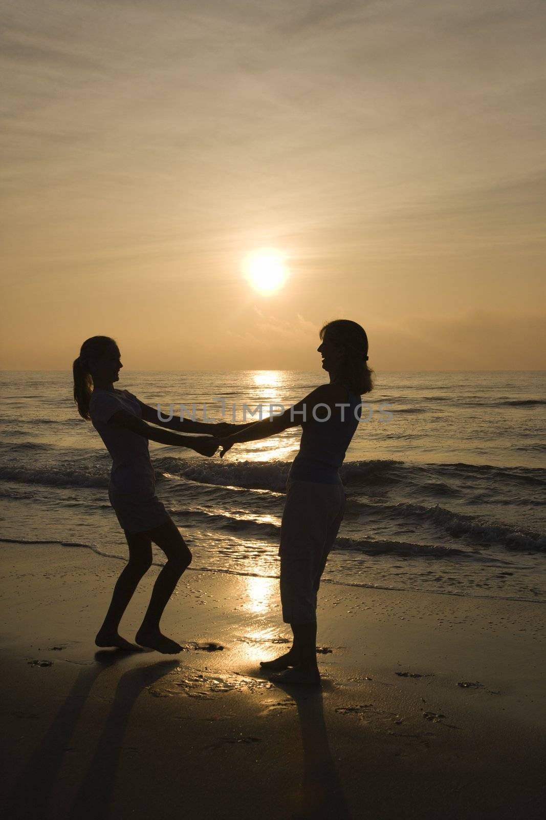 Caucasian mid-adult mother and teenage daughter holding hands spinngin on beach at sunset.