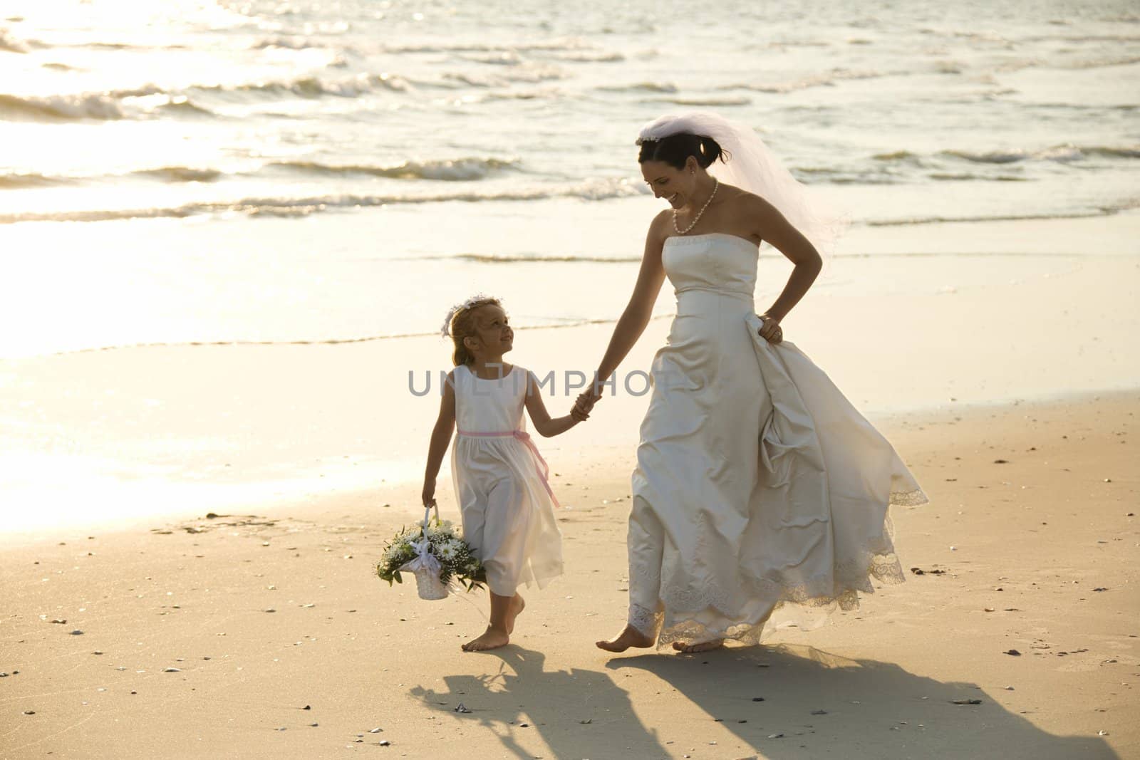 Caucasian mid-adult bride and flower girl holding hands walking barefoot on beach.