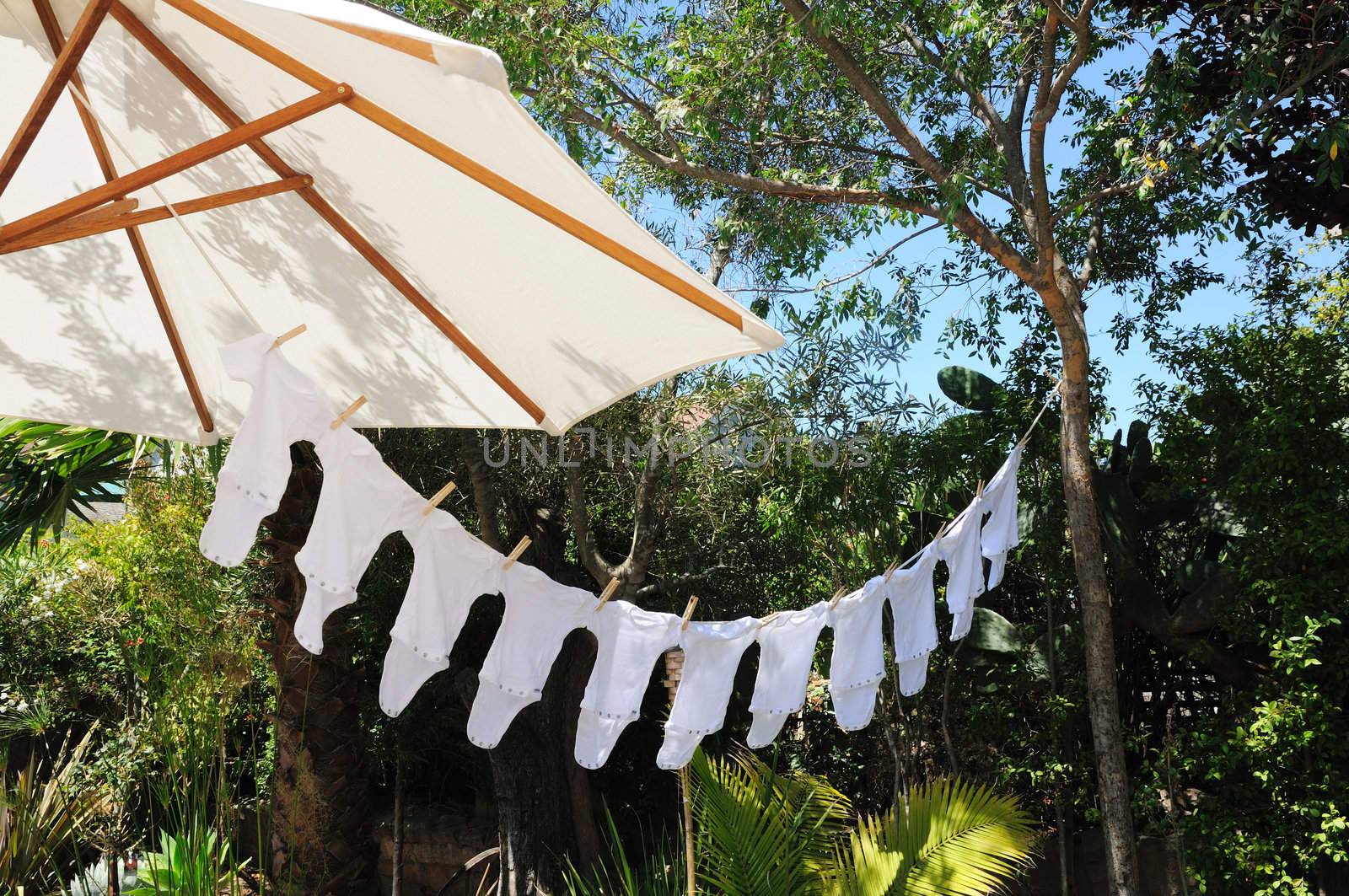 A row of baby onesies hanging from a clothesline suspended from a sun umbrella.