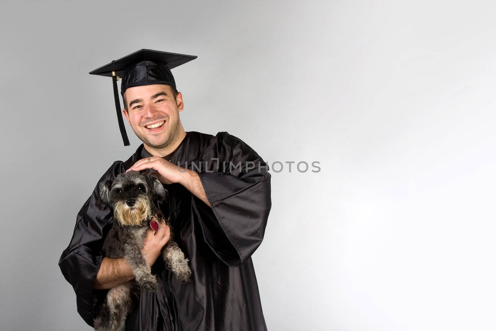 A recent college or high school graduate in his cap and gown holding his pet dog in his arms.