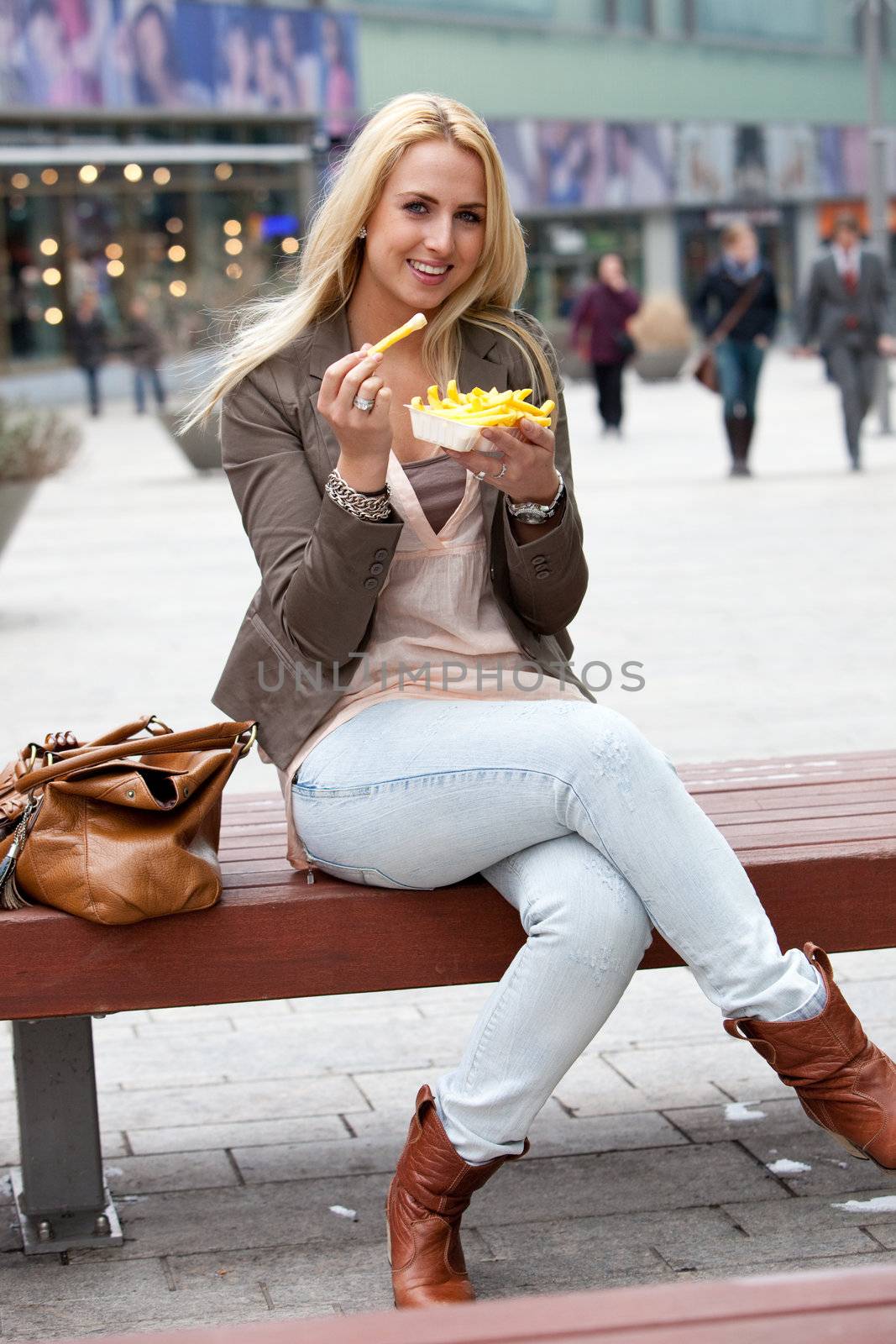 Pretty young blond girl sitting outdoors on a bench eating fries