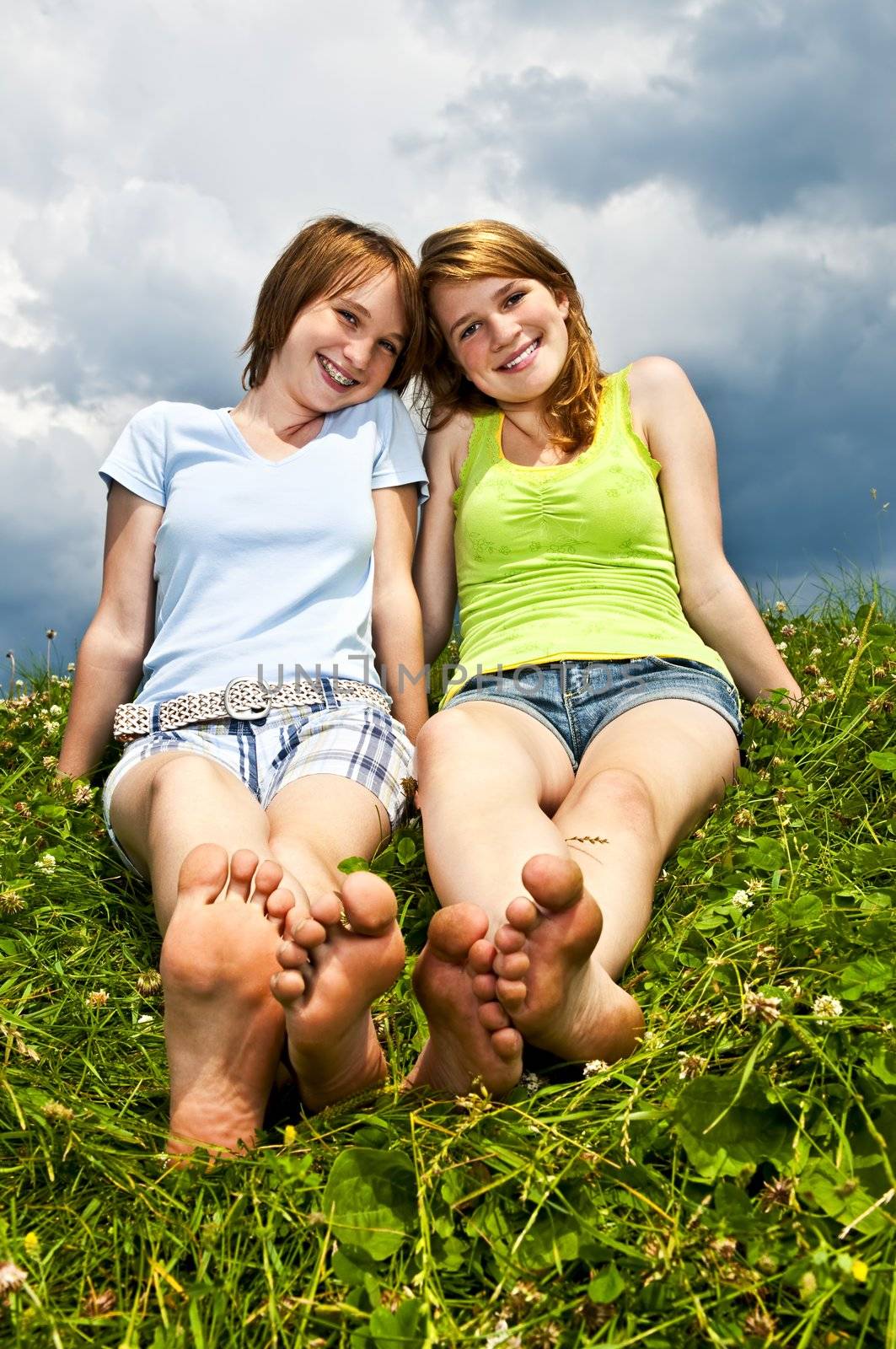 Two young teenage girl friends sitting barefoot on summer meadow