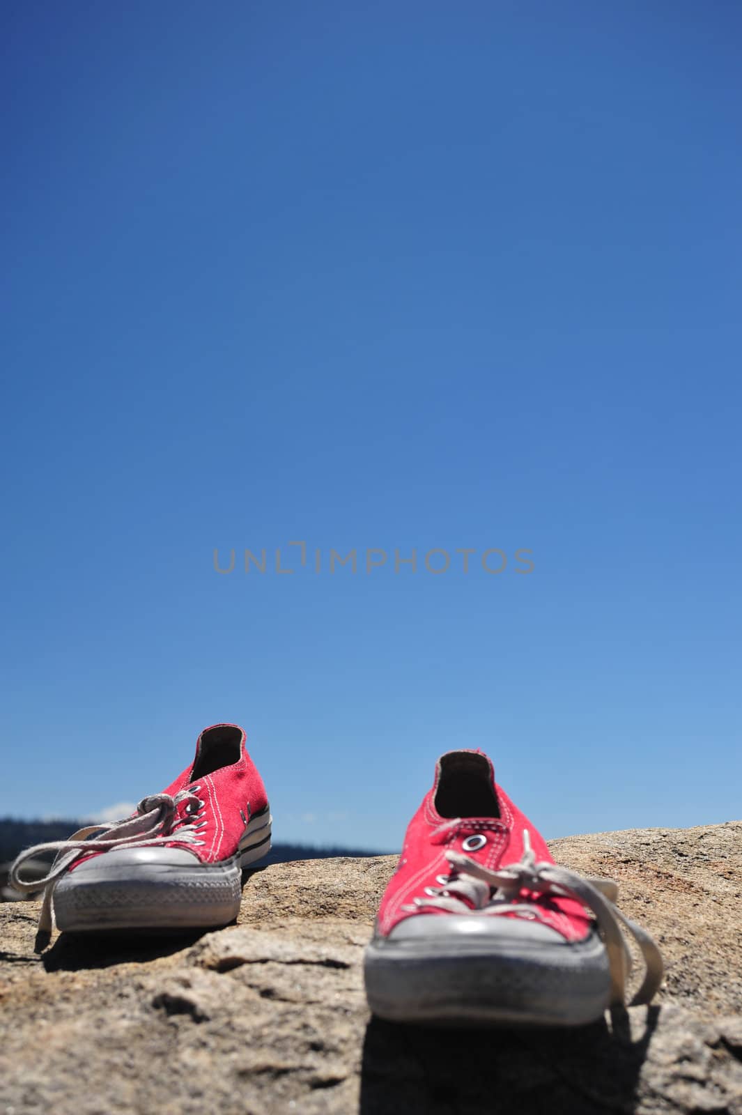 A pair of red tennis shoes by them selves on granite with a bright blue sky in the background.