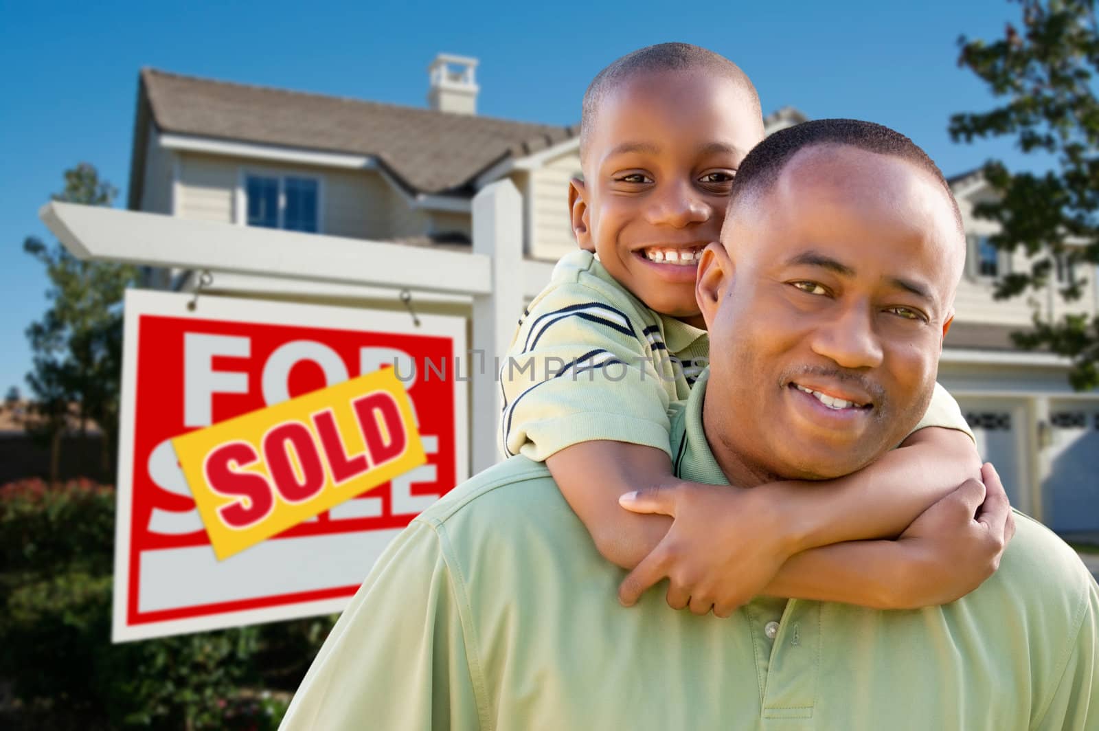 Happy African American Father and Son in Front of New Home and Real Estate Sign.