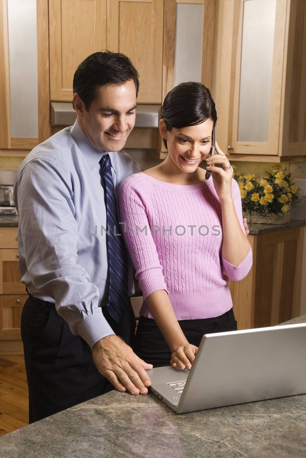 MId-adult female talking on phone and mid-adult male both looking at laptop computer in kitchen.