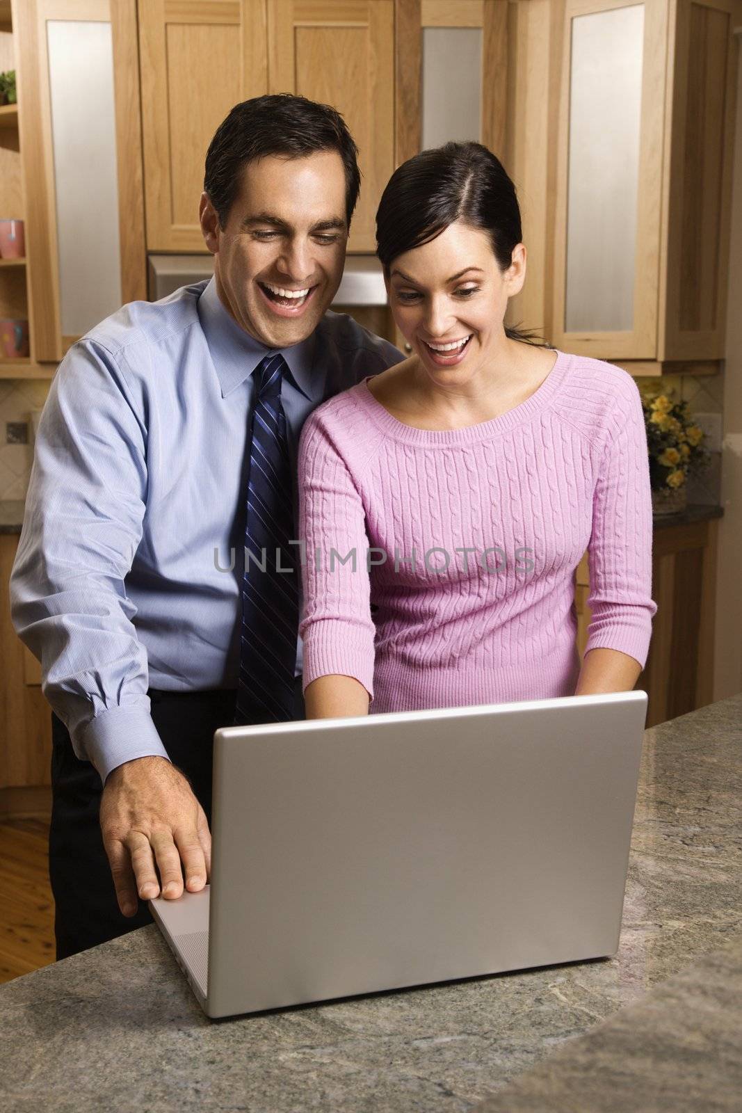 Mid-adult couple in kitchen looking and smiling at laptop computer.