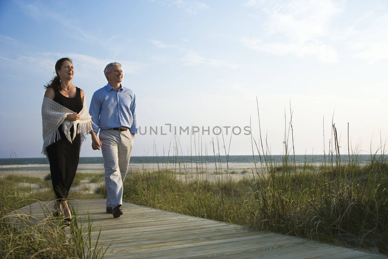 Caucasian mid-adult couple holding hands and walking down walkway at beach.