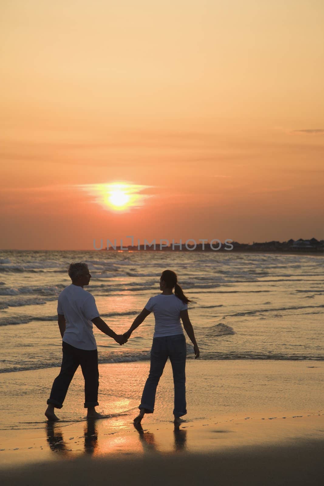 Mid-adult couple holding hands and walking on beach at sunset.