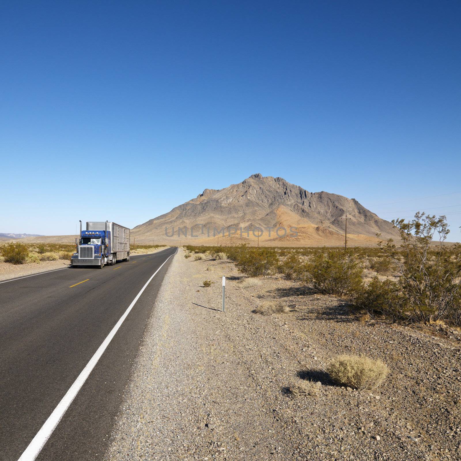 Tractor trailer driving on desert road with mountain in background.