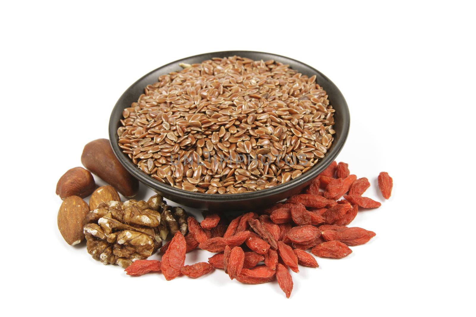 Brown linseed seeds in a small black bowl with mixed nuts and goji berries on a reflective white background