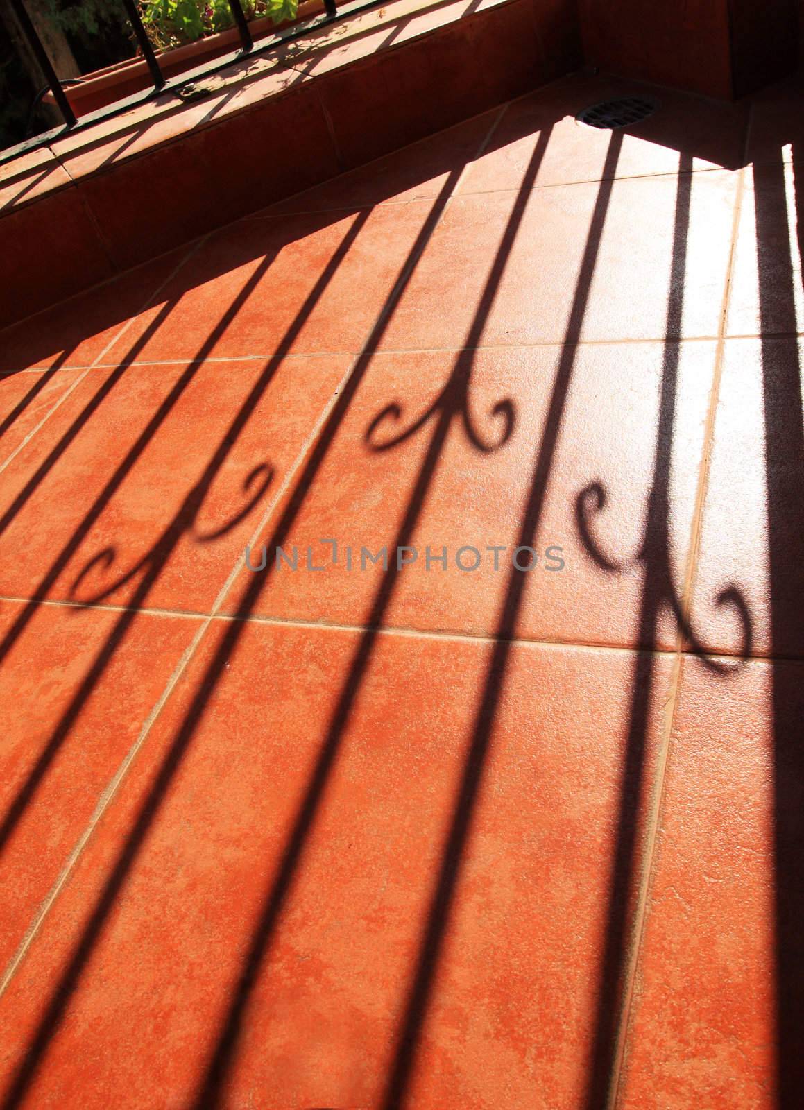 The grate shadow on a ceramic tiled balcony floor.Tiles are of bright terracota color .Shadow-pattern  looks geometric,   sharp enough and simetrical.