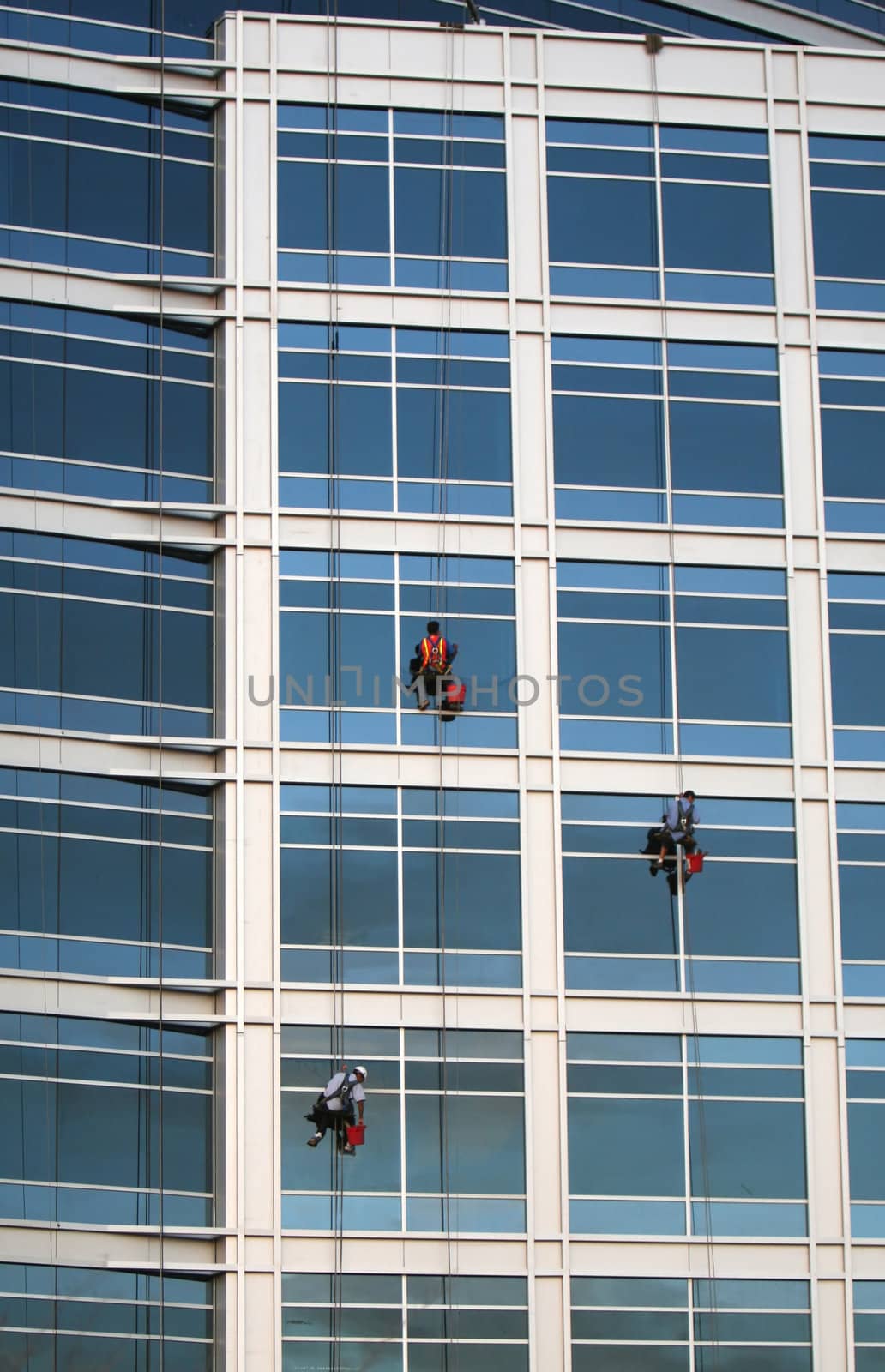 Three window washers descend on ropes high above the city. The building is a very modern glass structure.
