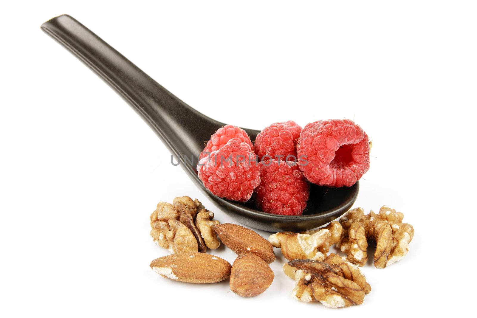 Red ripe frozen raspberries on a small black spoon with mixed nuts on a reflective white background