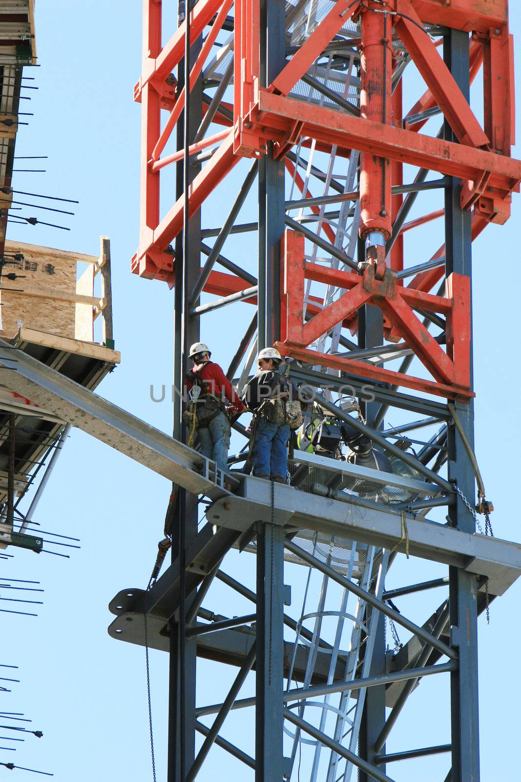 Two construction workers are tethered to the side of a crane that is being erected
